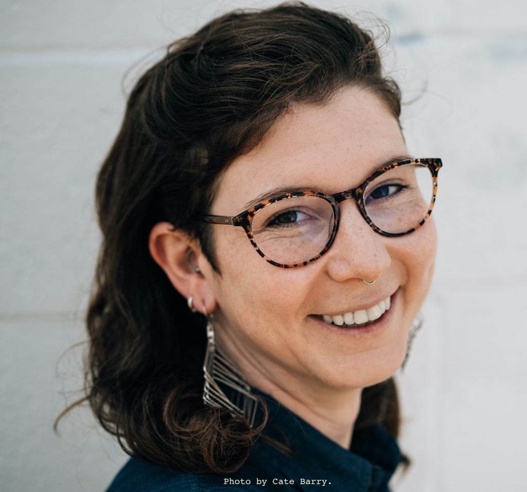 A headshot of a light-skinned, freckled femme with curly brown shoulder-length hair and brown eyes looking over their shoulder and smiling against a white wall. They are wearing a navy blue collar shirt, tortoiseshell glasses, and geometric silver earrings. They have silver rings in their septum, tragus, and ear lobe.